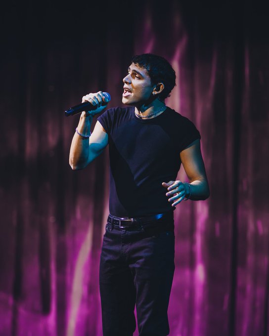 A young man with short dark hair stands on a stage holding a microphone close to his mouth as he sings with an open expressive face. He wears a black sleeveless t-shirt that shows his arms a thin necklace and dark pants with a belt. His right arm is raised slightly and he has a bracelet on his wrist. The background features a dark purple curtain.