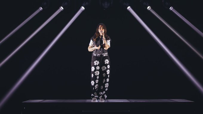 A young woman with long dark hair stands on a dark stage wearing a light-colored short-sleeve top black pants with white floral patterns and sneakers holding a microphone in her right hand with her left hand in her pocket illuminated by multiple purple spotlights beaming down from above creating beams of light on the floor.