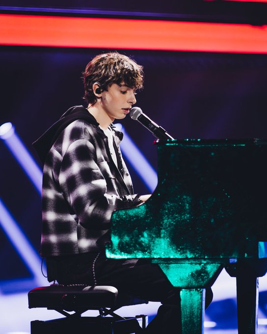 Young man with curly brown hair wearing a black plaid hoodie over a white shirt sits on a stool at a green grand piano on a stage with a microphone in hand, under bright stage lights with red and blue glowing backdrop elements.