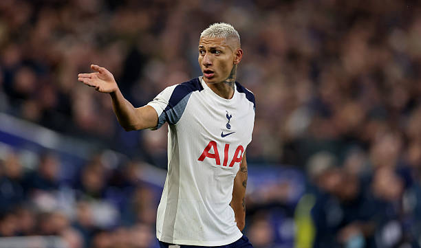 Brazilian soccer player Richarlison with platinum blonde hair stands on a stadium field wearing Tottenham Hotspur home kit featuring navy blue shoulders white body and Nike swoosh gesturing with right hand extended palm up toward blurred crowd and stadium seating in background during match.