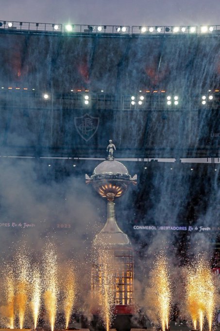 A large silver trophy cup with intricate engravings sits on a pedestal in a dimly lit stadium under floodlights and rainy atmosphere with the River Plate club emblem visible on the stadium structure behind it surrounded by colorful fireworks exploding in the background.