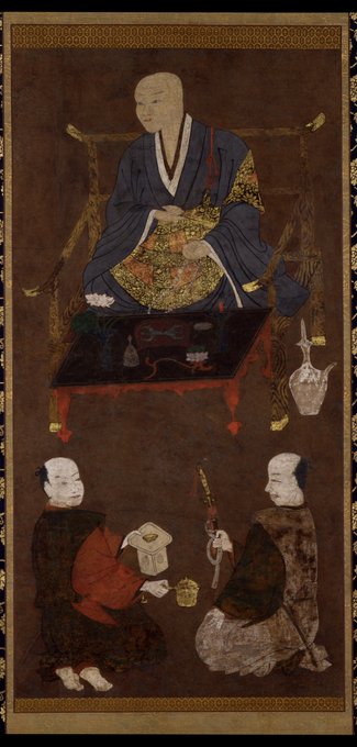 A traditional Japanese painting on silk with gold leaf details shows a bald central figure in blue and gold monk robes seated on a chair holding a fan in his right hand and prayer beads in his left, with two attendant figures in red and brown robes kneeling below him one holding a document and the other a staff, all set against a dark background with a low table in front featuring incense burner and vase.