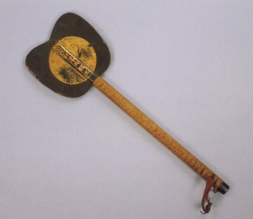 First image shows a wooden military fan with a dark brown oval-shaped blade featuring a central gold foil sun disk with intricate gold patterns and surrounding red ink inscriptions of deity names attached to a long light brown woven handle ending in a red leather strap. Second image is a close-up of the fan blade displaying the gold sun disk with Sanskrit-like characters in the center and additional red ink writings on the surrounding dark areas with visible wear and patina.
