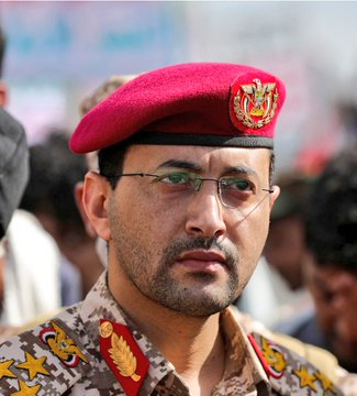 Three men wearing traditional Arab attire and headscarves stand in front of the tall Burj Khalifa skyscraper under a clear sky with clouds, one man raises his right hand while all hold rifles, surrounded by modern buildings and water. A separate image shows a man in military uniform with red beret and gold insignia on shoulder epaulets, standing among a crowd.