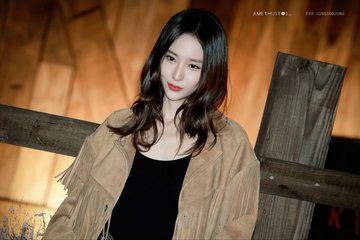 A woman with long wavy dark hair and red lips stands in a warmly lit wooden interior wearing a fringed tan suede jacket over a black tank top paired with jeans and a large silver belt buckle. Multiple similar shots show her posing against wooden beams and structures emphasizing the outfit details and neutral expression. Watermarks AMETHYSTOS and for Jung Soojung appear on each image.