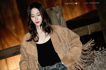 A woman with long wavy dark hair and red lips stands in a warmly lit wooden interior wearing a fringed tan suede jacket over a black tank top paired with jeans and a large silver belt buckle. Multiple similar shots show her posing against wooden beams and structures emphasizing the outfit details and neutral expression. Watermarks AMETHYSTOS and for Jung Soojung appear on each image.