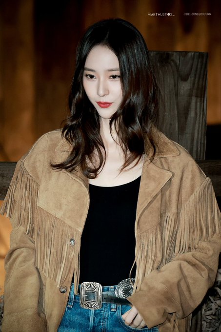 A woman with long wavy dark hair and red lips stands in a warmly lit wooden interior wearing a fringed tan suede jacket over a black tank top paired with jeans and a large silver belt buckle. Multiple similar shots show her posing against wooden beams and structures emphasizing the outfit details and neutral expression. Watermarks AMETHYSTOS and for Jung Soojung appear on each image.