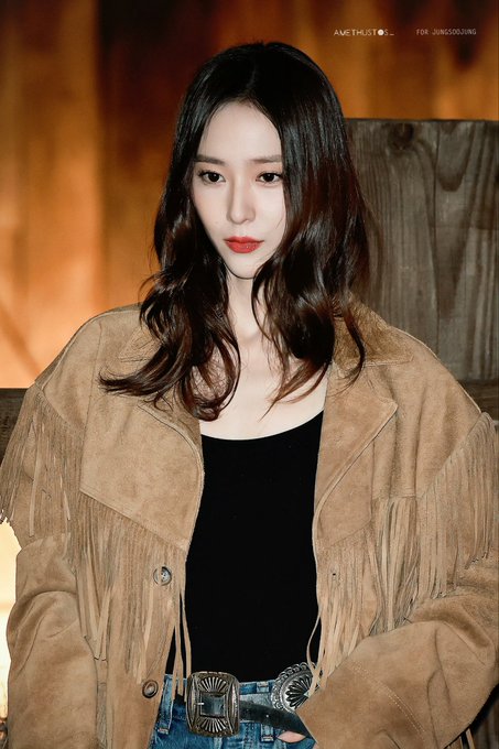 A woman with long wavy dark hair and red lips stands in a warmly lit wooden interior wearing a fringed tan suede jacket over a black tank top paired with jeans and a large silver belt buckle. Multiple similar shots show her posing against wooden beams and structures emphasizing the outfit details and neutral expression. Watermarks AMETHYSTOS and for Jung Soojung appear on each image.