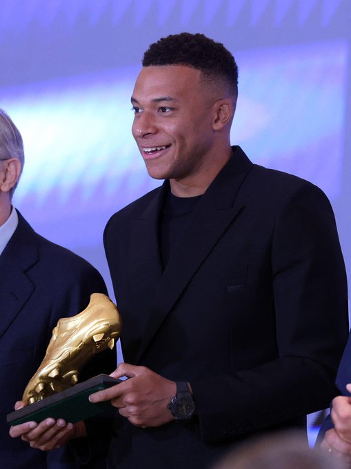 Kylian Mbappé stands on a stage holding a large golden boot trophy with a green base, dressed in a black suit and smiling broadly. An older man in a gray suit stands beside him on the left, also smiling. Another person in a suit is partially visible on the right. The background features blue lighting and a blurred audience.