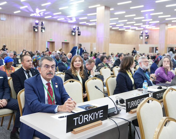 First image shows a formal conference room with wooden paneling and lighting fixtures where diverse international delegates in business suits sit around a table. A man in a blue suit with red tie and nameplate Turkiye gestures while seated next to another delegate. Nameplates indicate Turkiye and other countries on the table with microphones water bottles and documents. Second image depicts a large blue screen displaying UNESCO General Conference 42nd session logo and a live video feed of speakers including a man in suit labeled Turkiye. Rows of delegates from countries like France and Malta sit at tables facing the screen in a spacious hall with blue chairs. Third image captures delegates at a long table in a lit conference space with microphones documents and laptops. A man in blue suit with red tie types on a laptop while a woman in yellow blouse sits nearby. Various attendees in suits headscarves and traditional attire engage in discussions amid cameras and flags.