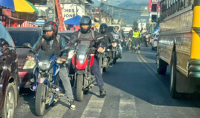 Multiple motorcycles and motorbikes parked or stopped in a line on a street with several riders wearing helmets and casual clothing some standing beside their vehicles a yellow school bus visible on the right side cars parked nearby and buildings with signs along the roadside in an urban setting with mountains in the background