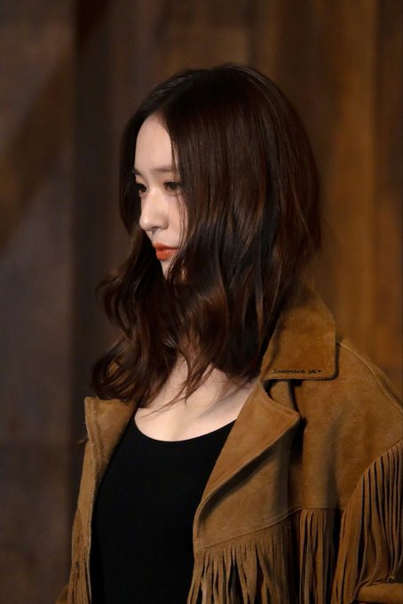 A woman with long wavy brown hair and light makeup stands in profile view wearing a brown fringed suede jacket over a black top in a dimly lit wooden interior setting. The jacket features fringe details on the sleeves and hem. Another similar image shows the same woman from a slightly different angle with the same outfit and setting.