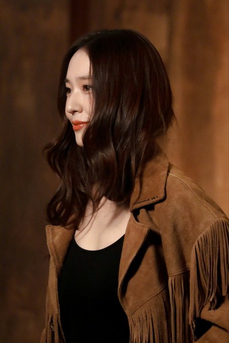 A woman with long wavy brown hair and light makeup stands in profile view wearing a brown fringed suede jacket over a black top in a dimly lit wooden interior setting. The jacket features fringe details on the sleeves and hem. Another similar image shows the same woman from a slightly different angle with the same outfit and setting.