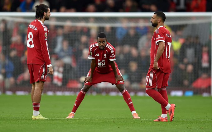 Three male soccer players in red Liverpool FC kits with white accents stand on a green grass field near a white goal net during a match. The player on the left has long dark hair in a ponytail wears number 8 jersey and red shorts with green socks. The central player in white kit with red accents squats with hands on knees. The right player with short dark hair wears number 17 jersey and red shorts with orange cleats. Background shows stadium seating with blurred spectators.