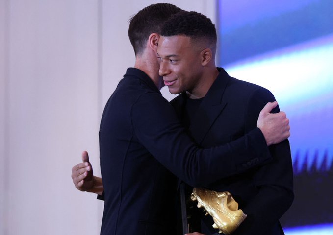 Two men stand on a stage embracing, the one on the left with short dark hair wearing a black suit and smiling, the one on the right with a fade haircut in a black suit holding a golden boot trophy with spikes, blue lighting in the background and a white wall.