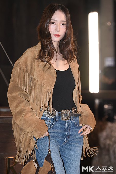 A woman with long wavy brown hair wearing a brown fringed suede jacket over a black tank top blue jeans with a silver belt and brown bag stands in a dimly lit indoor setting with background lights and blurred figures. In another image she waves with her right hand while dressed similarly accessorized with black boots and a round brown bag against a backdrop with grass-like elements and Louis Vuitton signage. Close-up shows her face with subtle makeup smiling softly. Fourth image captures her waving hand extended in the fringed jacket outfit with event lighting.