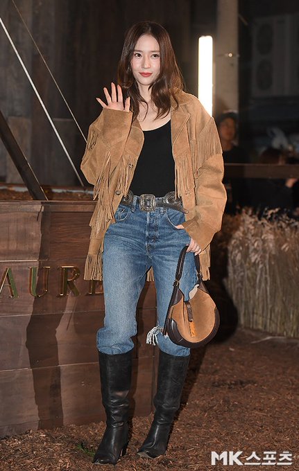 A woman with long wavy brown hair wearing a brown fringed suede jacket over a black tank top blue jeans with a silver belt and brown bag stands in a dimly lit indoor setting with background lights and blurred figures. In another image she waves with her right hand while dressed similarly accessorized with black boots and a round brown bag against a backdrop with grass-like elements and Louis Vuitton signage. Close-up shows her face with subtle makeup smiling softly. Fourth image captures her waving hand extended in the fringed jacket outfit with event lighting.