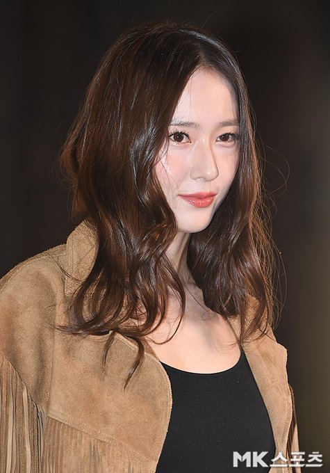 A woman with long wavy brown hair wearing a brown fringed suede jacket over a black tank top blue jeans with a silver belt and brown bag stands in a dimly lit indoor setting with background lights and blurred figures. In another image she waves with her right hand while dressed similarly accessorized with black boots and a round brown bag against a backdrop with grass-like elements and Louis Vuitton signage. Close-up shows her face with subtle makeup smiling softly. Fourth image captures her waving hand extended in the fringed jacket outfit with event lighting.