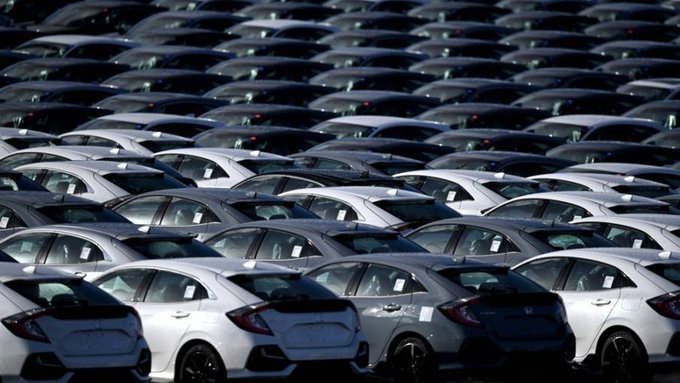 Portrait of Xi Jinping standing in front of Chinese national flags, wearing a dark suit with white shirt and blue tie, smiling slightly. Aerial view of numerous white and gray Honda Civic cars parked in rows at a dealership lot under daylight.