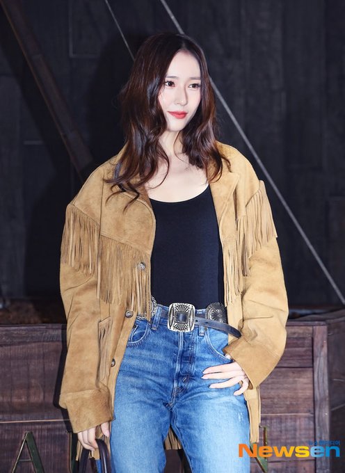 A woman with long wavy dark hair stands in a dark indoor setting with wooden structures behind her. She wears a long tan fringed suede jacket over a black tank top tucked into high-waisted blue jeans fastened with a wide brown belt featuring a large silver rectangular buckle. She holds a small black handbag in her right hand and stands with her weight on one leg posing confidently.