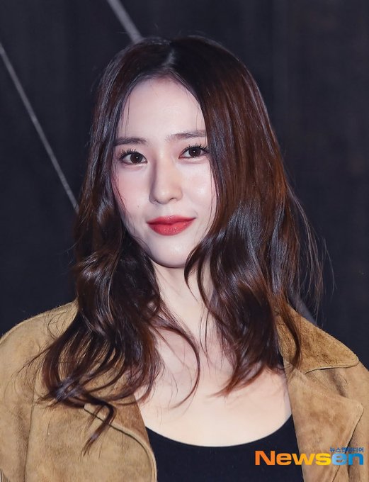 A young woman with long wavy brown hair stands outdoors at night wearing a fringed tan suede jacket over a black tank top, blue ripped jeans, a brown leather belt, and tall black boots, holding a brown leather handbag, posing with hands on hips near wooden barriers and Christmas lights in the background. In the second image, she raises her right hand in a peace sign gesture, smiling slightly with red lipstick, her hair cascading over the suede jacket. The third image shows a close-up of her face and upper body, with brown hair framing her features, red lips, and the suede jacket visible, set against a dark backdrop.