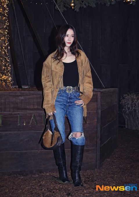 A young woman with long wavy brown hair stands outdoors at night wearing a fringed tan suede jacket over a black tank top, blue ripped jeans, a brown leather belt, and tall black boots, holding a brown leather handbag, posing with hands on hips near wooden barriers and Christmas lights in the background. In the second image, she raises her right hand in a peace sign gesture, smiling slightly with red lipstick, her hair cascading over the suede jacket. The third image shows a close-up of her face and upper body, with brown hair framing her features, red lips, and the suede jacket visible, set against a dark backdrop.
