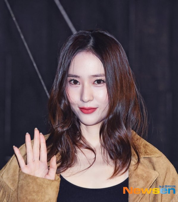 A young woman with long wavy brown hair stands outdoors at night wearing a fringed tan suede jacket over a black tank top, blue ripped jeans, a brown leather belt, and tall black boots, holding a brown leather handbag, posing with hands on hips near wooden barriers and Christmas lights in the background. In the second image, she raises her right hand in a peace sign gesture, smiling slightly with red lipstick, her hair cascading over the suede jacket. The third image shows a close-up of her face and upper body, with brown hair framing her features, red lips, and the suede jacket visible, set against a dark backdrop.