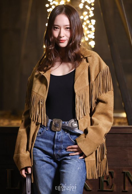 A woman with long wavy dark hair stands smiling in a dimly lit setting with hanging lights and wooden elements. She wears a fringed tan suede jacket over a black tank top, blue jeans, and holds a bag, with Ralph Lauren text visible on a wooden sign behind her. In another view, she poses similarly with the jacket details prominent. A third image shows her from a side angle wearing tall black boots, ripped jeans, and the same outfit against a rustic backdrop with lights.