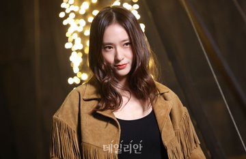 A woman with long wavy dark hair stands smiling in a dimly lit setting with hanging lights and wooden elements. She wears a fringed tan suede jacket over a black tank top, blue jeans, and holds a bag, with Ralph Lauren text visible on a wooden sign behind her. In another view, she poses similarly with the jacket details prominent. A third image shows her from a side angle wearing tall black boots, ripped jeans, and the same outfit against a rustic backdrop with lights.