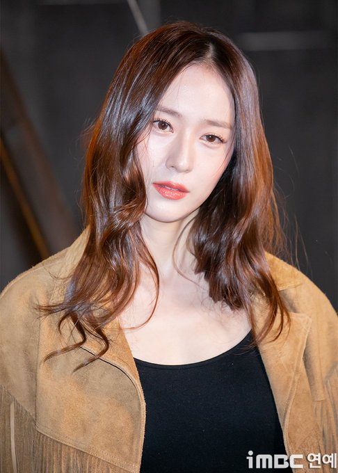 Portrait of a woman with long wavy brown hair parted in the middle, fair skin, almond-shaped eyes with subtle eyeliner, thin lips with light pink lipstick, neutral expression, wearing a black sleeveless top under an open fringed brown suede jacket, standing indoors against a dark blurred background.