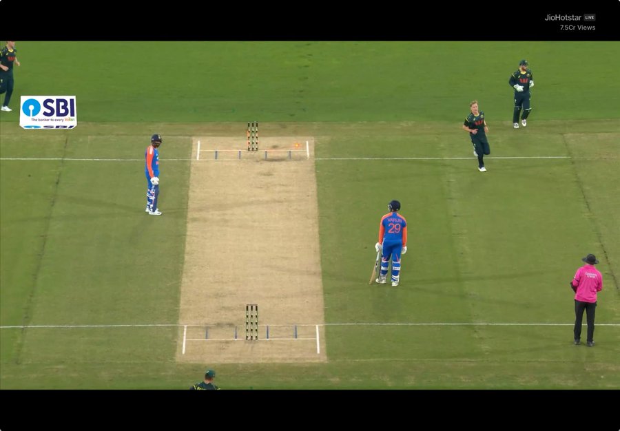 Cricket field with green grass and a brown pitch in the center. Players in blue uniforms including a batsman holding a bat near the crease and a fielder in green nearby. Umpires in white and pink attire stand on the sides. Sponsor boards like SBI visible on the boundary. Additional fielders positioned around the outfield.