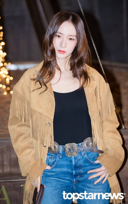 A young woman with long wavy brown hair stands posing with hands on hips at an outdoor event area at night illuminated by string lights and a bridge structure in the background she wears a fringed beige suede jacket over a black tank top high-waisted blue jeans with a large silver belt buckle and holds a small brown handbag the image includes watermarks from topstarnews