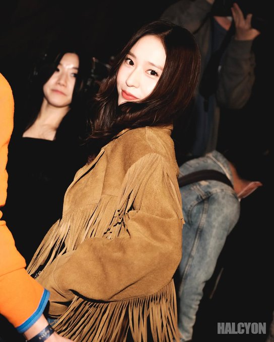 First image shows a woman with long dark hair smiling while wearing a tan fringed suede jacket over a black top standing in a dimly lit indoor space with blurred people in background including one in orange top. Second image depicts the same woman posing with hand on hip wearing the tan fringed suede jacket over black tank top and blue jeans with belt near string lights and grass in a nighttime outdoor setting.