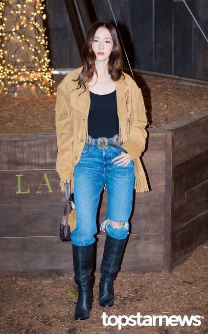 Woman with long wavy brown hair and subtle makeup stands in a three-quarter pose, wearing a long beige fringed suede jacket over a black tank top, high-waisted distressed blue jeans with rips at knees, a wide brown leather belt with large buckle, black over-the-knee boots, and holding a small tan handbag with fringe details. Background includes warm holiday string lights, a dark wooden elevated platform, and a sign with Ralph Lauren logo. Watermarks from topstarnews appear at bottom corners.
