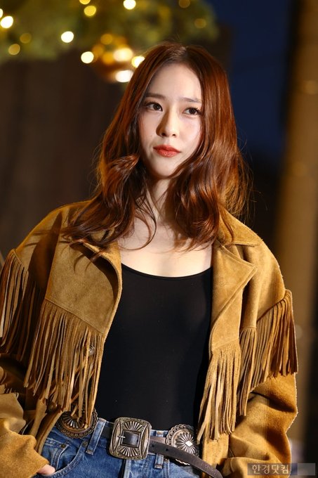 A woman with long wavy brown hair wearing a fringed tan suede jacket over a black tank top and blue jeans stands in a nighttime outdoor setting with golden hanging lights and a decorated Christmas tree in the background. She poses with hands in pockets looking at the camera. In another image she waves with her right hand smiling. Additional similar poses show her from different angles emphasizing the outfit details like the belt and fringes.