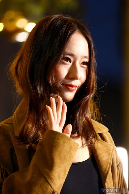 A woman with long wavy brown hair wearing a fringed tan suede jacket over a black tank top and blue jeans stands in a nighttime outdoor setting with golden hanging lights and a decorated Christmas tree in the background. She poses with hands in pockets looking at the camera. In another image she waves with her right hand smiling. Additional similar poses show her from different angles emphasizing the outfit details like the belt and fringes.