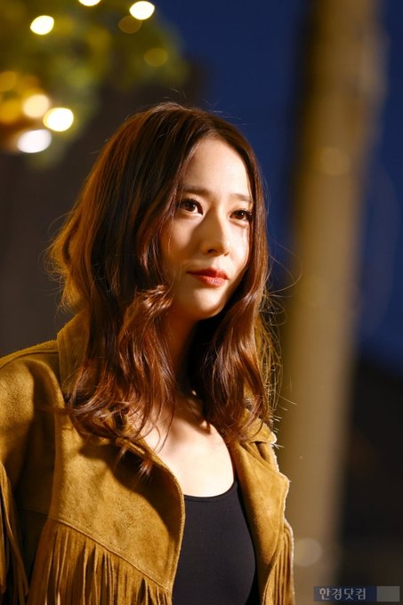 A woman with long wavy brown hair wearing a fringed tan suede jacket over a black tank top and blue jeans stands in a nighttime outdoor setting with golden hanging lights and a decorated Christmas tree in the background. She poses with hands in pockets looking at the camera. In another image she waves with her right hand smiling. Additional similar poses show her from different angles emphasizing the outfit details like the belt and fringes.