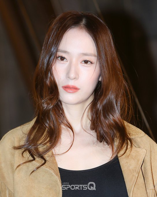 Woman with long wavy brown hair parted in middle smiling slightly at camera wearing black sleeveless top under open beige coat standing against dark background in fashion event setting.