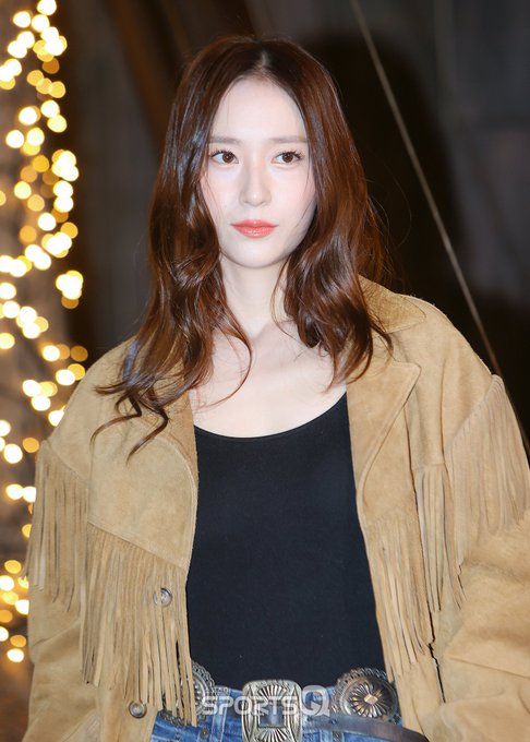 A woman with long wavy brown hair stands smiling at the camera wearing a fringed tan suede jacket over a black tank top blue jeans with rips at the knees and a wide belt with silver concho details holding a brown bag with fringe in front of a wooden backdrop labeled SPOTV and surrounded by sparkling string lights at night. Another image shows the same woman from a closer angle with a neutral expression highlighting her outfit details including the jackets fringe and the belts design against the same festive background.