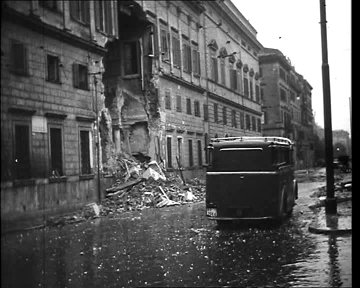 Black-and-white photograph depicts a street scene in Rome with the British Embassy building severely damaged, showing collapsed sections of the facade, exposed interiors, rubble scattered on the wet pavement, and a vintage car parked nearby under a streetlamp.