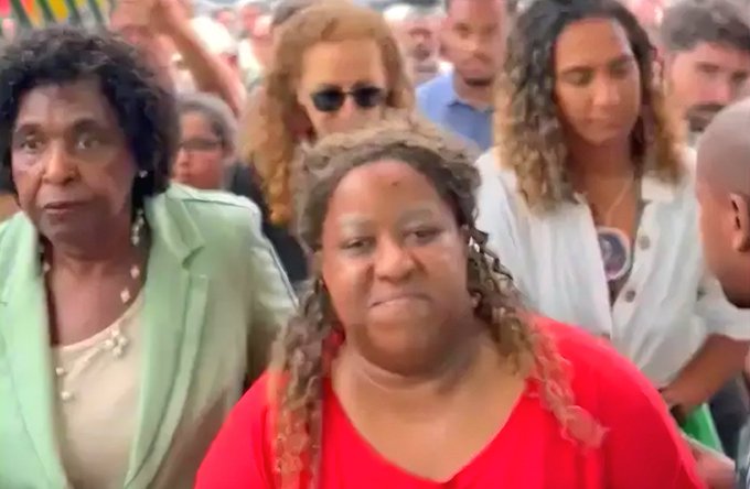 A group of diverse individuals stands closely in an outdoor crowd setting. Several women with curly hair wear casual outfits including a green jacket over a white top an older woman in green with earrings and a red-dressed woman with necklace. Men in the background wear sunglasses and light clothing. Some arms are raised suggesting excitement or gesturing. The scene appears lively with neutral expressions on faces.
