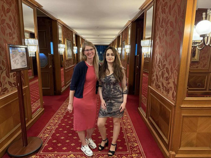 First image shows two women standing in a luxurious hotel hallway with wooden paneling red carpet ornate wall decor and chandeliers. One woman wears glasses a navy cardigan and red dress with white sneakers the other has long dark hair a patterned dress and high heels. Second image depicts a conference stage with a large screen displaying Sol 2022 logo Earth image and names Dr Beatriz Villarroel and Colonel Karl Nell. Two people sit in chairs on stage one woman with long hair in casual attire and one man in suit next to a microphone stand.