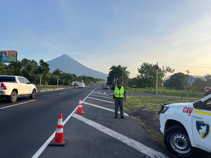 A highway scene with white and silver vehicles parked on the roadside including a marked white patrol vehicle with CIV logo and shield emblem. Orange traffic cones are placed along the road edges. A person in a high-visibility green vest and helmet stands directing traffic. Palm trees line the background with a prominent conical volcano visible in the distance under a clear sky.