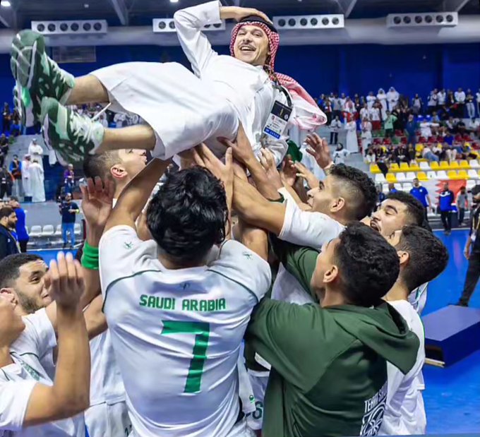 Group of young Saudi male athletes in white thobes and green jerseys with number 7 and Saudi Arabia text, wearing red and white shemaghs, joyfully lifting one smiling player in mid-air with arms raised in celebration inside a blue-seated indoor sports arena with green podium.