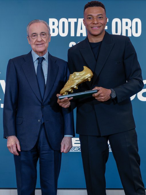 An older man in a dark blue suit with tie stands next to a younger man in a black suit, both smiling and holding a large golden boot trophy on a blue background with European shoe branding and text.
