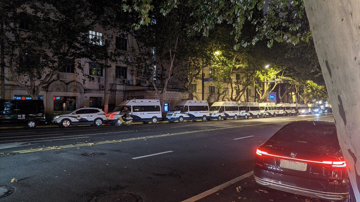 Nighttime urban street scene with multiple white police vans and vehicles parked in a line along the roadside illuminated by overhead yellow streetlights. Surrounding buildings with arched windows and trees with leaves visible in the background. A black sedan with red taillights positioned in the foreground on the wet pavement. Additional police cars and vans clustered near a building entrance suggesting heightened security presence.