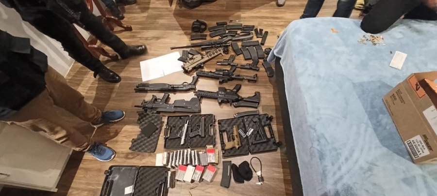 First image shows multiple rifles, handguns, magazines, and cases scattered on wooden floor near blue bed and cardboard boxes in indoor room with people in background. Second image displays handguns, magazines, cases, backpack, and orange box on blue bed with white pillows and lamp nearby. Third image depicts rifles, shotgun, magazines, and backpack laid out on wooden floor in indoor setting with shoes visible.