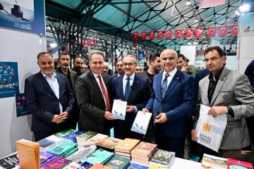 First image shows a large group of officials and attendees in formal attire standing together cutting a red ribbon at the entrance of a spacious tent venue decorated with numerous Turkish flags hanging from the ceiling and walls, with a prominent banner reading Malatya Anadolu Kitap Fuarı Bir Kitap in the background. Second image depicts several men in suits gathered around a display table covered with various colorful books, some holding books with blue covers, positioned in front of a booth with digital screens and promotional materials. Third image features a diverse group of people including men and women in business attire standing behind a table laden with stacks of books at a booth labeled with red banners for Milli Egitim Malatya Valiligi Mudurlugu, surrounded by educational signage. Fourth image captures an elderly woman in a dark hijab and long coat standing with a man in a blue suit and glasses, accompanied by a young girl in a white dress and three young boys in casual clothes holding items, all posing in front of bookshelves in an exhibition hall setting.