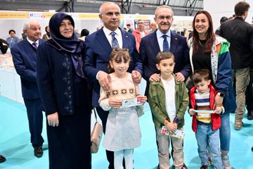 First image shows a large group of officials and attendees in formal attire standing together cutting a red ribbon at the entrance of a spacious tent venue decorated with numerous Turkish flags hanging from the ceiling and walls, with a prominent banner reading Malatya Anadolu Kitap Fuarı Bir Kitap in the background. Second image depicts several men in suits gathered around a display table covered with various colorful books, some holding books with blue covers, positioned in front of a booth with digital screens and promotional materials. Third image features a diverse group of people including men and women in business attire standing behind a table laden with stacks of books at a booth labeled with red banners for Milli Egitim Malatya Valiligi Mudurlugu, surrounded by educational signage. Fourth image captures an elderly woman in a dark hijab and long coat standing with a man in a blue suit and glasses, accompanied by a young girl in a white dress and three young boys in casual clothes holding items, all posing in front of bookshelves in an exhibition hall setting.