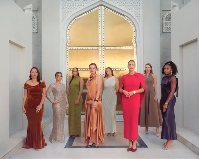 Six women standing in a row pose for a group photograph in a grand hall with white walls intricate golden arched doorways and decorative elements. They wear various elegant long dresses in colors including red orange green gray and purple some with off-shoulder or cape styles and high heels. Their expressions are confident and the setting appears formal and luxurious.